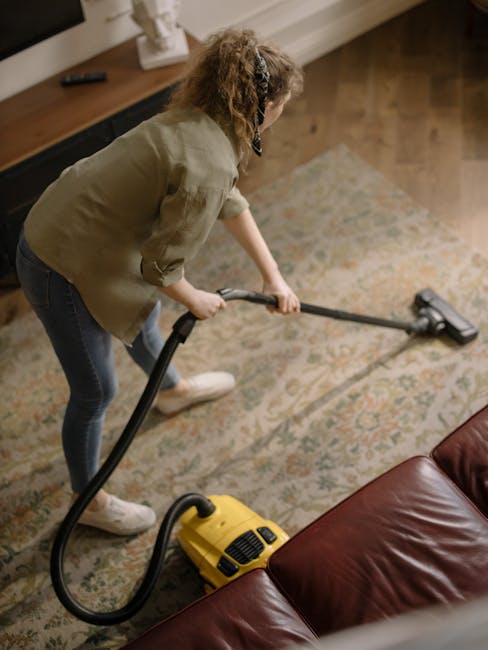 A close-up view of a canister vacuum cleaner with a transparent dust container filled with debris and bubbles, positioned on a pink carpeted floor in a residential room. The vacuum has a flexible, pat