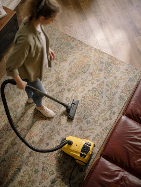 A professional cleaning specialist dressed in full protective gear, including a white suit, mask, and gloves, is performing deep cleaning using a fogging or steam disinfectant machine on a beige carpet in a modern living room. The room features a wooden shelving unit with decorative items, a large mirror, and potted plants, and is well-lit with natural light. The cleaning process appears thorough, with visible mist or vapor on the carpet, indicating sanitisation action. This image exemplifies surface cleaning and hygiene maintenance typical of services provided by Carpet Cleaning Ilford, as featured on the page titled 'Carpet cleaning Ilford Broadway flats and maisonettes' on the company's website.
