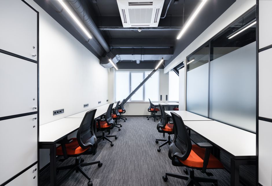 A person in casual work attire is performing surface cleaning indoors, using a long-handled mop or squeegee on a dark carpeted floor. The room appears to be a commercial space, possibly an office or c