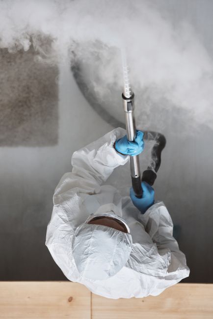 A professional cleaner dressed in full protective gear, including a white disposable coverall, gloves, mask, and shoe covers, is using a steam cleaning machine on a beige carpet in a bright, modern li