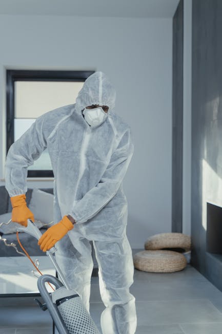 A person operating a black vacuum cleaner with a silver telescopic tube on a light wooden floor, cleaning a residential room. The individual is wearing dark shorts, white socks, and gray slippers, wit