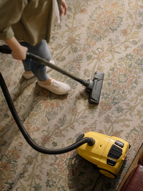 A person wearing beige shoes and blue jeans is operating a yellow canister vacuum cleaner with a black hose, using a cleaning head attachment to perform surface cleaning on a large, patterned area rug in a room. The rug features a floral design in muted colors, and the room is well-lit with natural or artificial light. The vacuum cleaner is positioned on the floor, and the person is holding the hose handle while working on the rug, indicating a deep cleaning process. This scene illustrates domestic cleaning practices aligned with professional standards maintained by Carpet Cleaning Ilford, focusing on ensuring hygienic and thoroughly cleaned surfaces.