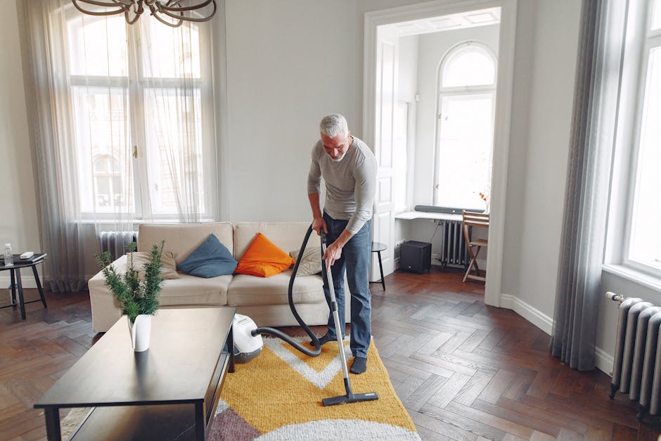 A middle-aged man with gray hair and glasses is performing surface cleaning in a well-lit living room, vacuuming a yellow and white patterned rug with a modern vacuum cleaner. The room features a light-colored sofa adorned with colorful cushions in blue, orange, and beige, and is decorated with sheer curtains and large windows allowing natural light to fill the space. A wooden coffee table with a potted plant is positioned near the sofa, and a small side table with a glass of water is visible. The wooden floor is clean and polished, reflecting the ambient light, and a small desk with a chair is situated in the corner. The scene emulates a professional domestic cleaning environment, highlighting the importance of maintaining hygiene and surface cleanliness with effective cleaning tools, as provided by Carpet Cleaning Ilford, specialists in home and commercial cleaning.