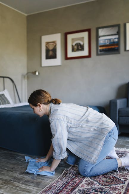 A person wearing beige shoes and blue jeans is operating a yellow canister vacuum cleaner with a black hose, using a cleaning head attachment to perform surface cleaning on a large, patterned area rug in a room. The rug features a floral design in muted colors, and the room is well-lit with natural or artificial light. The vacuum cleaner is positioned on the floor, and the person is holding the hose handle while working on the rug, indicating a deep cleaning process. This scene illustrates domestic cleaning practices aligned with professional standards maintained by Carpet Cleaning Ilford, focusing on ensuring hygienic and thoroughly cleaned surfaces.