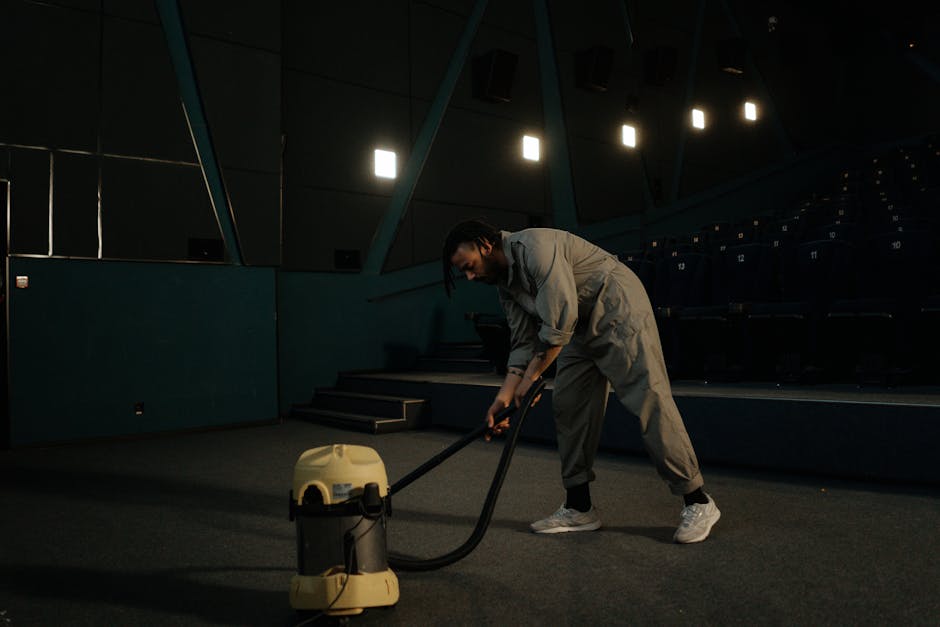 A person using a vacuum cleaner with a long, black hose attachment to deep clean a large traditional patterned area rug featuring floral motifs in beige, green, and orange tones. The individual is sta