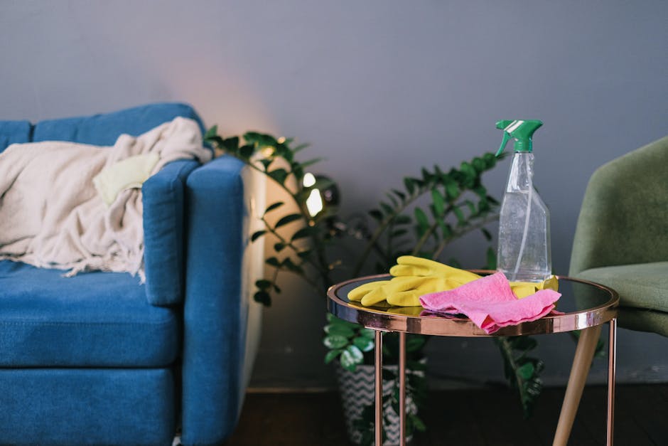 A close-up of a living room corner showing a blue fabric sofa with a light-colored blanket draped over its armrest. To the right, a small round side table with a reflective, dark surface holds yellow rubber cleaning gloves, a pink cloth, and a clear spray bottle with a green trigger, indicating cleaning supplies. Behind the table, a large potted plant with broad green leaves is visible, adding a touch of natural decor. The background features a plain gray wall with soft ambient lighting, highlighting the cleanliness and tidy arrangement of the area, typical of a space maintained through surface cleaning and domestic sanitisation practices by Carpet Cleaning Ilford.