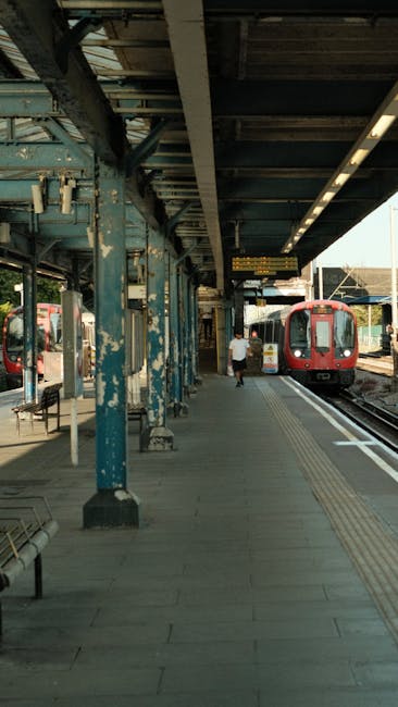 A clean, well-maintained train station platform at Ilford station showing a tiled concrete surface free of dirt or litter. There are metal benches along the platform, with some painted in a weathered blue finish. The platform is covered by a metal canopy supported by blue painted columns, which show signs of peeling paint and rust. Under the canopy, bright lighting fixtures illuminate the area. A waiting person and a red train car are visible in the background, with the train partially on the tracks. The scene demonstrates a hygienic environment suitable for passengers, reflecting the high standards of surface cleaning and sanitisation typical of professional cleaning services like those provided by Carpet Cleaning Ilford.