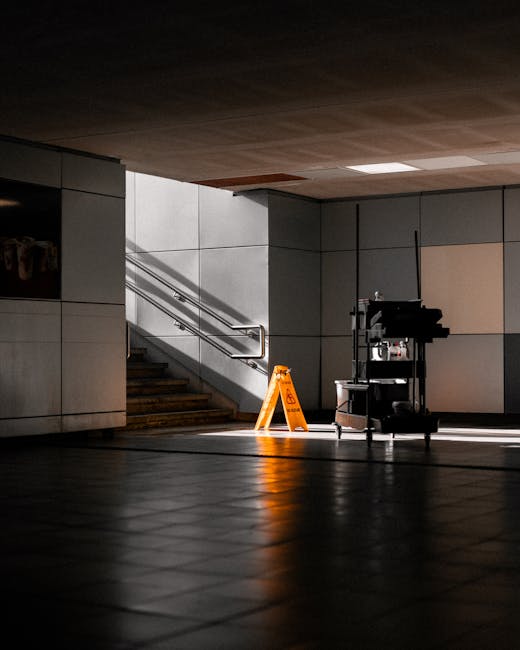 A modern, tiled indoor space featuring a staircase with metallic handrails leading upward, illuminated by natural light streaming in from an unseen window. The floor is composed of polished, dark tiles that reflect the light and shadows, creating a clean and shiny surface. Near the base of the stairs, there is an orange caution wet floor sign and a mobile cleaning cart equipped with various cleaning supplies and tools. The walls are constructed of large, smooth, light-colored panels, and the ceiling appears to be wooden with built-in lighting fixtures. The scene depicts a well-maintained, hygienic environment suitable for surface cleaning and deep cleaning services promoted by Carpet Cleaning Ilford, emphasizing sanitisation and upkeep of commercial or residential spaces.
