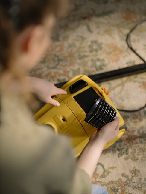 Close-up view of a person operating a yellow portable carpet cleaning or drying machine, held with both hands, inside a room with a floral-patterned carpet. The image showcases the person's blurred profile with focusing on the machine's air vent and control panel, emphasizing the surface cleaning and drying process. The room's natural lighting highlights the carpet's detailed pattern and the cleanliness after professional deep cleaning by Carpet Cleaning Ilford, as referenced on the webpage titled 'Valentines Park rug cleaning before and after case study'. The setting suggests ongoing maintenance or post-cleaning sanitisation to restore the carpet's appearance and hygiene.