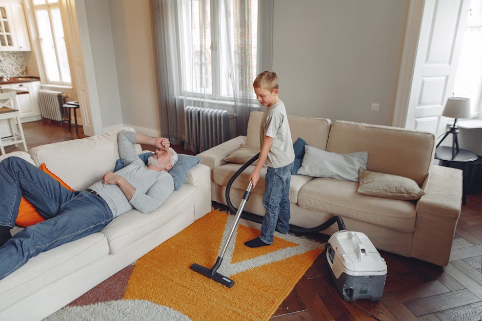 A young boy is vacuuming a carpet in a bright, modern living room with a grey sofa and beige armchair. The room features large windows with grey curtains, allowing natural light to illuminate the space. The floor is a dark wood, partially covered by an orange and beige patterned rug. An older man with grey hair and beard is lying on the sofa, resting with one arm behind his head and a relaxed expression. The vacuum cleaner, placed on the floor, is connected to the boy, who is focused on deep cleaning the carpet. The room is clean and tidy, with neutral-colored cushions on the sofa, a side table with a lamp, and a radiator visible in the background. Carpet Cleaning Ilford offers professional surface cleaning services, including rug deep cleaning, as demonstrated in this scene highlighting domestic cleaning steps for maintaining hygiene and cleanliness.